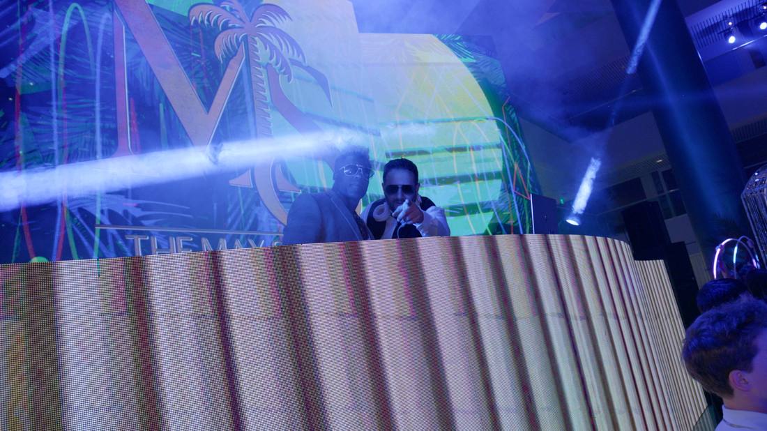 Two guests posing at the DJ booth beneath a palm-themed event sign