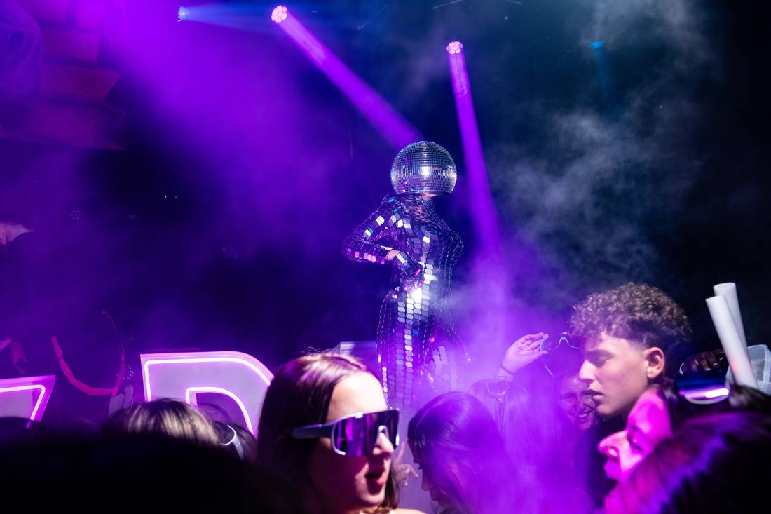 Performer in a mirrored disco-ball costume dancing above the crowd under purple light