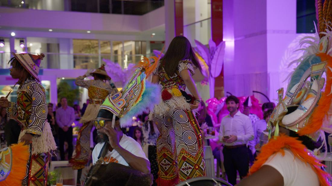 Carnival-costumed performers in feathered headdresses dancing indoors